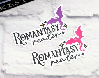May include: Two stickers with the text "Romantasy reader" and a dragon illustration. One sticker has a purple dragon, the other a pink dragon. Both have yellow star accents. The stickers are on a white brick background.