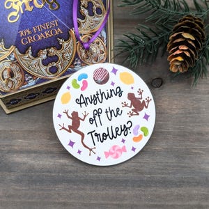 May include: A white circular ornament with the text "Anything off the Trolley?" features colorful candy and two brown frog illustrations. A purple ribbon and a decorative box with gold accents are visible in the background.