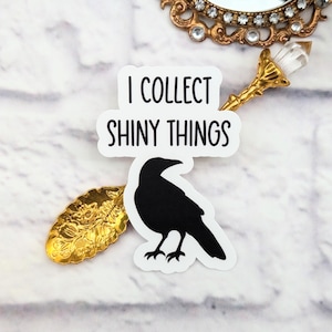 Puede incluir: Una pegatina blanca con el texto "I COLLECT SHINY THINGS" sobre una silueta de cuervo negro. La pegatina está junto a una cuchara decorativa dorada y un espejo dorado ornamentado.