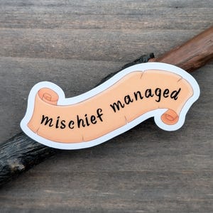 May include: A sticker featuring the phrase "mischief managed" in black script on a light orange scroll. The scroll has a white border and is set against a dark wooden background. A dark brown, textured stick is also visible.