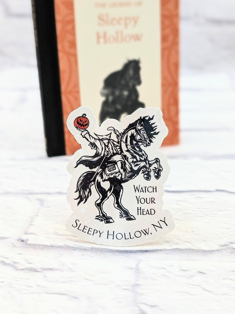 Pode incluir: Um adesivo preto e branco do Cavaleiro Sem Cabe&ccedil;a montando um cavalo com uma ab&oacute;bora esculpida na m&atilde;o. O adesivo diz "Watch Your Head" e "Sleepy Hollow, NY".