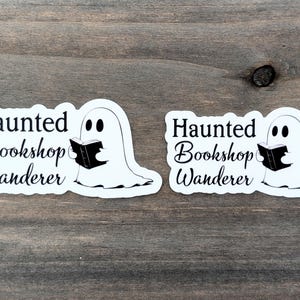 May include: Two white stickers featuring a ghost reading a book. The stickers have the text "Haunted Bookshop Wanderer" in black script and a ghost illustration. They are on a wooden surface.