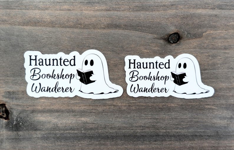 May include: Two white stickers featuring a ghost reading a book. The stickers have the text "Haunted Bookshop Wanderer" in black script and a ghost illustration. They are on a wooden surface.