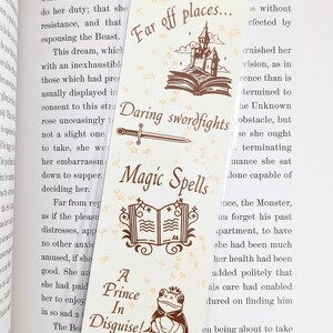 May include: A cream-colored bookmark with fantasy-themed illustrations and text. The bookmark features a castle, a sword, an open book, and a frog prince. Text includes "Far off places...", "Daring swordfights", "Magic Spells", and "A Prince In Disguise!"