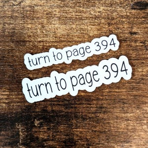 Könnte beinhalten: Zwei weiße Aufkleber mit schwarzem Text auf einer braunen Holzoberfläche. Der Text auf den Aufklebern lautet "drehen Sie zur Seite 394".