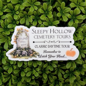 Pode incluir: Um autocolante branco com o texto "Sleepy Hollow Cemetery Tours" e um gráfico de lápide. O autocolante inclui o texto "Classic Daytime Tour" e uma ilustração de abóbora. A lápide diz "Irving 1783-1859".