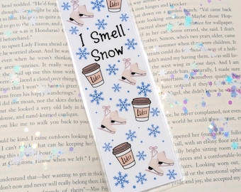Puede incluir: Marcapáginas transparente con la frase "I Smell Snow" e ilustraciones de patines de hielo, copos de nieve y tazas de café. Las tazas de café tienen la palabra "Lukes". El marcapáginas está sobre un libro abierto.