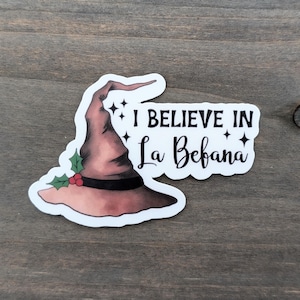 May include: A sticker featuring a witch's hat in shades of brown and pink, adorned with holly and berries. The text "I BELIEVE IN La Befana" is displayed in black, with star accents. The sticker is on a wooden surface.