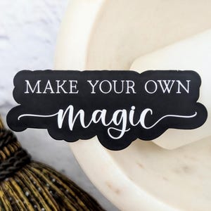 May include: A black sticker with the words "MAKE YOUR OWN magic" in white script. The sticker is on a white surface, with a broom and a mortar and pestle in the background.
