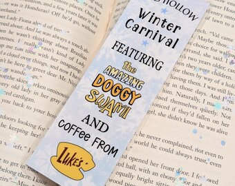 Puede incluir: Un marcapáginas con texto del Carnaval de Invierno de Stars Hollow, con "The Amazing Doggy Swami" y "Coffee from Luke's". El marcapáginas es azul claro con diseños de estrellas y copos de nieve, y está colocado en un libro abierto.