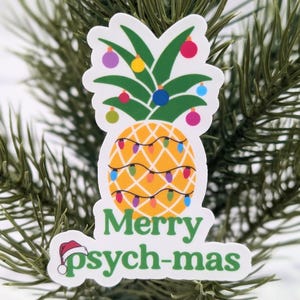 May include: A white sticker featuring a pineapple adorned with colourful baubles and fairy lights. The text "Merry Psych-mas" is written beneath the pineapple.