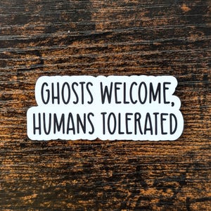 May include: White sticker with black text that reads "Ghosts Welcome, Humans Tolerated" on a dark brown wooden background.