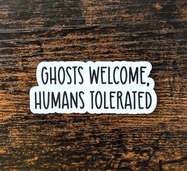 May include: White sticker with black text that reads "Ghosts Welcome, Humans Tolerated" on a dark brown wooden background.