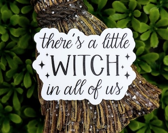 May include: A decorative sticker with the text "there's a little WITCH in all of us" in black script. The sticker is white with a decorative border and is placed on a miniature broom. The broom is brown and gold.