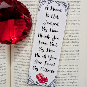 Könnte beinhalten: Ein Lesezeichen mit weißem Hintergrund und schwarzem Text, der lautet: "Ein Herz wird nicht danach beurteilt, wie sehr man liebt. Sondern danach, wie sehr man geliebt wird.". Das Lesezeichen zeigt ein Paar rote Rubinschuhe mit weißem Hintergrund und roten Schleifen.