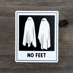 Pode incluir: Adesivo retangular com duas figuras de fantasmas brancos sobre fundo preto. O texto "NO FEET" está impresso em branco na parte inferior. O adesivo tem uma borda branca e está sobre uma superfície de madeira.