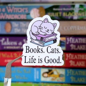 Może przedstawiać: Biała naklejka z ilustracją śpiącego kota na dwóch książkach. Naklejka zawiera czarny tekst "Books. Cats. Life is Good.". Tło przedstawia rozmyty stos książek.