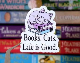 Könnte beinhalten: Ein weißer Aufkleber mit einer Illustration einer schlafenden Katze auf zwei Büchern. Der Aufkleber hat den Text "Books. Cats. Life is Good." in Schwarz. Der Hintergrund zeigt einen unscharfen Stapel von Büchern.