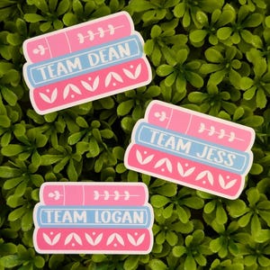 Puede incluir: Tres pegatinas con un diseño de pila de libros. Cada pegatina tiene una combinación de colores rosa y azul con las palabras "TEAM DEAN", "TEAM JESS" y "TEAM LOGAN" impresas. Las pegatinas están sobre un fondo verde y frondoso.