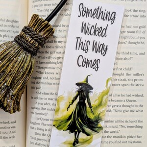 Puede incluir: Un marcapáginas con una bruja con un sombrero puntiagudo y un vestido verde, caminando hacia afuera de la vista del espectador. El fondo es una acuarela amarilla y verde. El texto "Something Wicked This Way Comes" está impreso en negro en el marcapáginas.