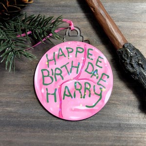 Może przedstawiać: Różowa, okrągła ozdoba świąteczna z napisem "HAPPEE BIRTH DAE HARRY" w kolorze zielonym. Posiada srebrny metalowy wierzch i różową wstążkę. Ozdobne gwiazdki są rozrzucone po powierzchni.