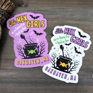 Puede incluir: Dos pegatinas coloridas con temática de brujas. Las pegatinas presentan el texto "The Hex Girls" y "We're Gonna Put a Spell on You" con un caldero, murciélagos y notas musicales. También se incluye el nombre de la ciudad "Oakhaven, MA".
