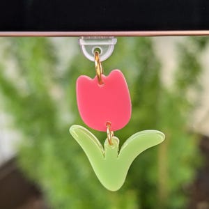 May include: A pair of tulip-shaped earrings. The earrings feature a bright pink flower and light green leaves, connected by gold-colored hoops. The earrings are hanging in front of a blurred green background.