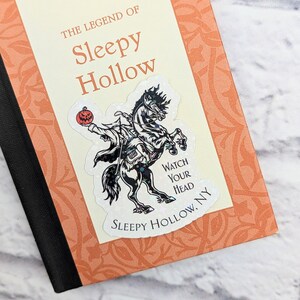 Pode incluir: Um adesivo com uma ilustra&ccedil;&atilde;o em preto e branco do Cavaleiro Sem Cabe&ccedil;a montando um cavalo. O adesivo est&aacute; em um livro intitulado "A Lenda de Sleepy Hollow" de Washington Irving. O adesivo diz "Watch Your Head Sleepy Hollow, NY".
