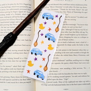 Könnte beinhalten: Ein skurriles Lesezeichen mit einem sich wiederholenden Muster aus hellblauen Autos, gelben Enten, orangefarbenen und lila Sternen und braunen Besen. Das Lesezeichen liegt auf einem Buch mit Text.