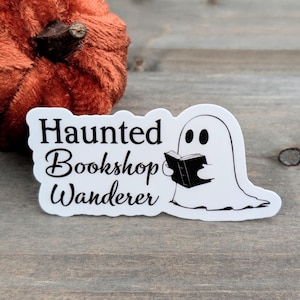 May include: A white sticker featuring the text "Haunted Bookshop Wanderer" and a cartoon ghost reading a book. The sticker is on a wooden surface, with a small orange pumpkin in the background.