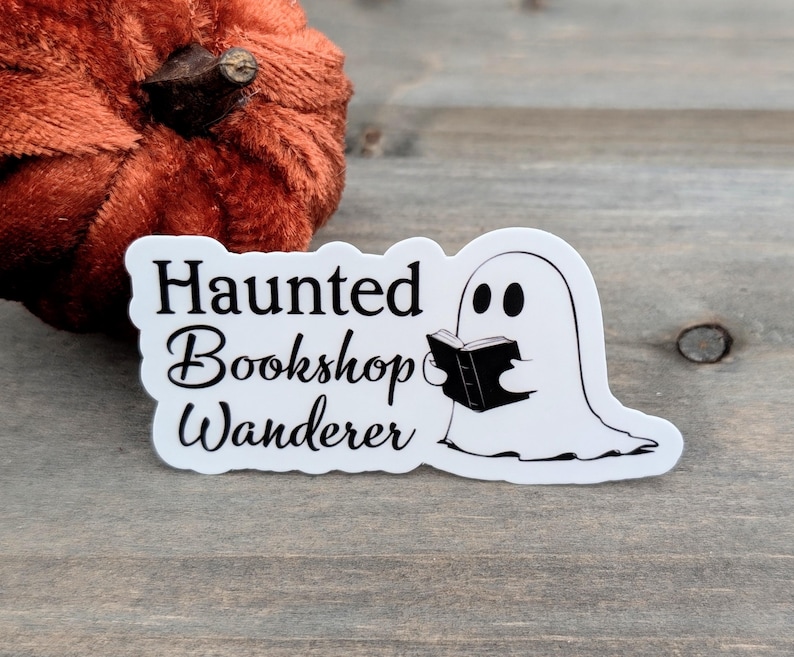 May include: A white sticker featuring the text "Haunted Bookshop Wanderer" and a cartoon ghost reading a book. The sticker is on a wooden surface, with a small orange pumpkin in the background.