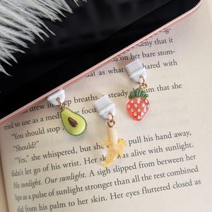 May include: Three fruit-shaped phone charger port charms: a green avocado, a yellow banana, and a red strawberry. Each charm has a white plug and a gold-colored loop for attachment. The charms are on a book.