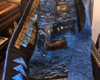 May include: A blue and brown quilt featuring a cabin scene. The cabin is lit up with yellow lights and there is a boat in the water in front of it. The quilt is folded and sitting on a brown leather chair.