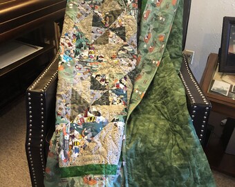 May include: A green and brown patchwork quilt with a Peanuts camping theme. The quilt is folded and draped over a brown leather chair.