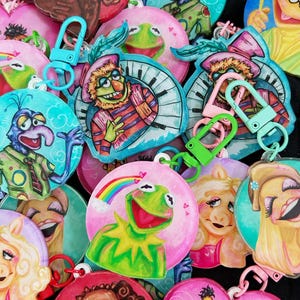 May include: A collection of colorful, round keychains featuring various Muppet characters. Each keychain has a unique design with vibrant colors and attached carabiner clips. The keychains showcase characters like Kermit the Frog, Miss Piggy, and others.