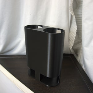 May include: A black plastic double cup holder with a rounded base. The holder is designed to hold two cups or glasses.