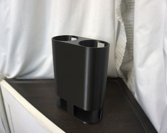 Op de afbeelding: Een zwarte dubbele bekerhouder van plastic met een afgeronde basis. De houder is ontworpen om twee bekers of glazen te houden.