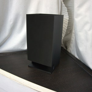 May include: A black rectangular speaker with a small base. The speaker is sitting on a dark brown surface.