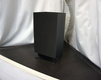 May include: A black rectangular speaker with a small base. The speaker is sitting on a dark brown surface.