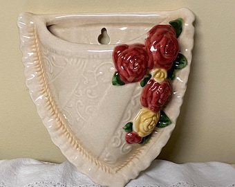May include: A white ceramic wall planter shaped like a heart with a scalloped edge. The planter has a raised floral design and features three red and yellow roses.