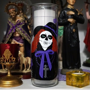 Puede incluir: Una vela de vidrio con un diseño negro y morado que presenta una calavera con cabello rojo y un lazo morado. La vela está rodeada de figuras religiosas.