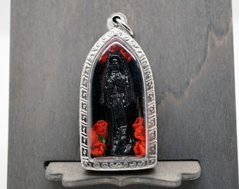 May include: A silver pendant with a black figure and red roses. The pendant has an arched shape with a decorative border and a loop for a chain. The background is black, set against a grey wooden stand.