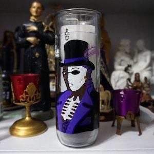 May include: A glass candle with a painted image of a person wearing a purple jacket, a black top hat with a feather, and a white eye patch. The person has a black and white face. The candle is on a white surface.