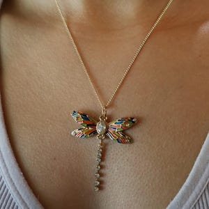 Puede incluir: Un collar de cadena dorada con un colgante de lib&eacute;lula colorido. La lib&eacute;lula tiene un cristal transparente en el centro y una fila de cristales transparentes que cuelgan de su cuerpo.