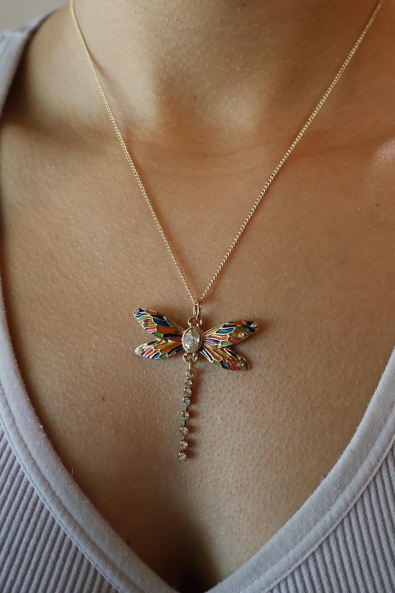 Puede incluir: Un collar de cadena dorada con un colgante de lib&eacute;lula colorido. La lib&eacute;lula tiene un cristal transparente en el centro y una fila de cristales transparentes que cuelgan de su cuerpo.