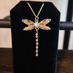 Puede incluir: Un collar de oro con un colgante de lib&eacute;lula colorido y una cadena de piedras de cristal transparentes.