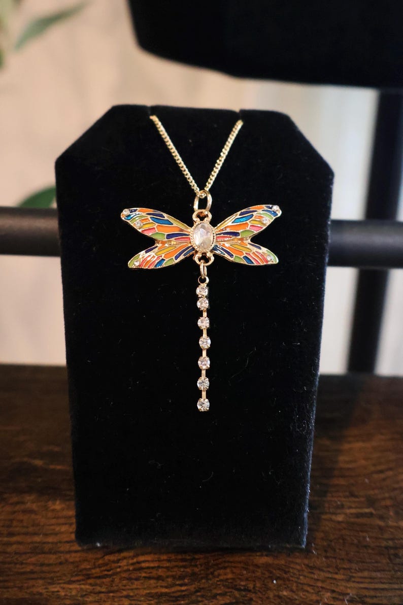 Puede incluir: Un collar de oro con un colgante de lib&eacute;lula colorido y una cadena de piedras de cristal transparentes.