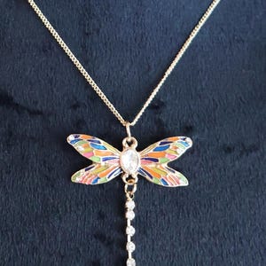 Puede incluir: Un collar de cadena dorada con un colgante de lib&eacute;lula colorido. La lib&eacute;lula tiene un cristal transparente en el centro y una cadena de cristales transparentes que cuelga.