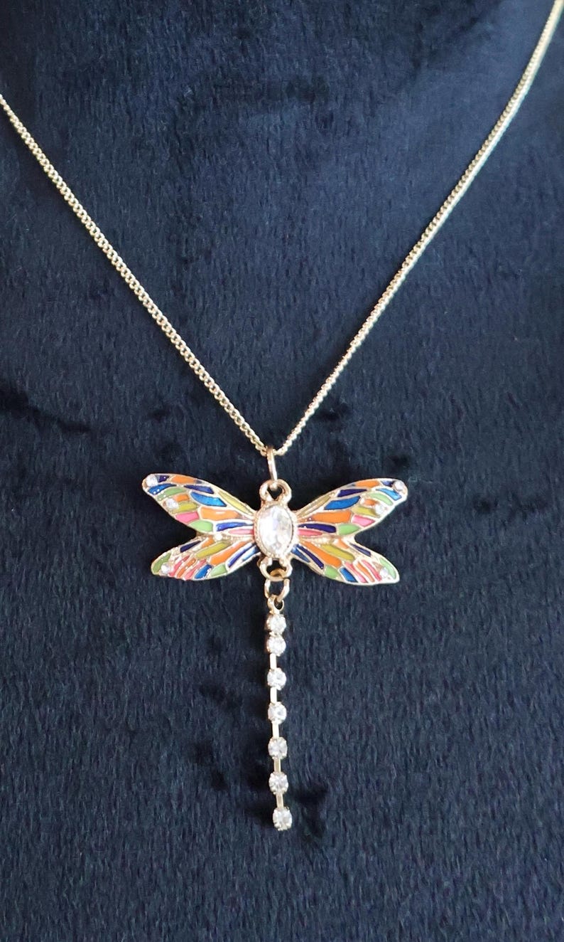 Puede incluir: Un collar de cadena dorada con un colgante de lib&eacute;lula colorido. La lib&eacute;lula tiene un cristal transparente en el centro y una cadena de cristales transparentes que cuelga.
