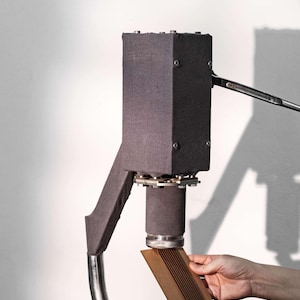 Op de afbeelding: Een donkergrijze industri&euml;le machine met een rechthoekige bovenkant en een gebogen metalen arm. Een persoon houdt een houten stuk onder de cilindrische basis van de machine. De machine heeft een complex mechanisch ontwerp.